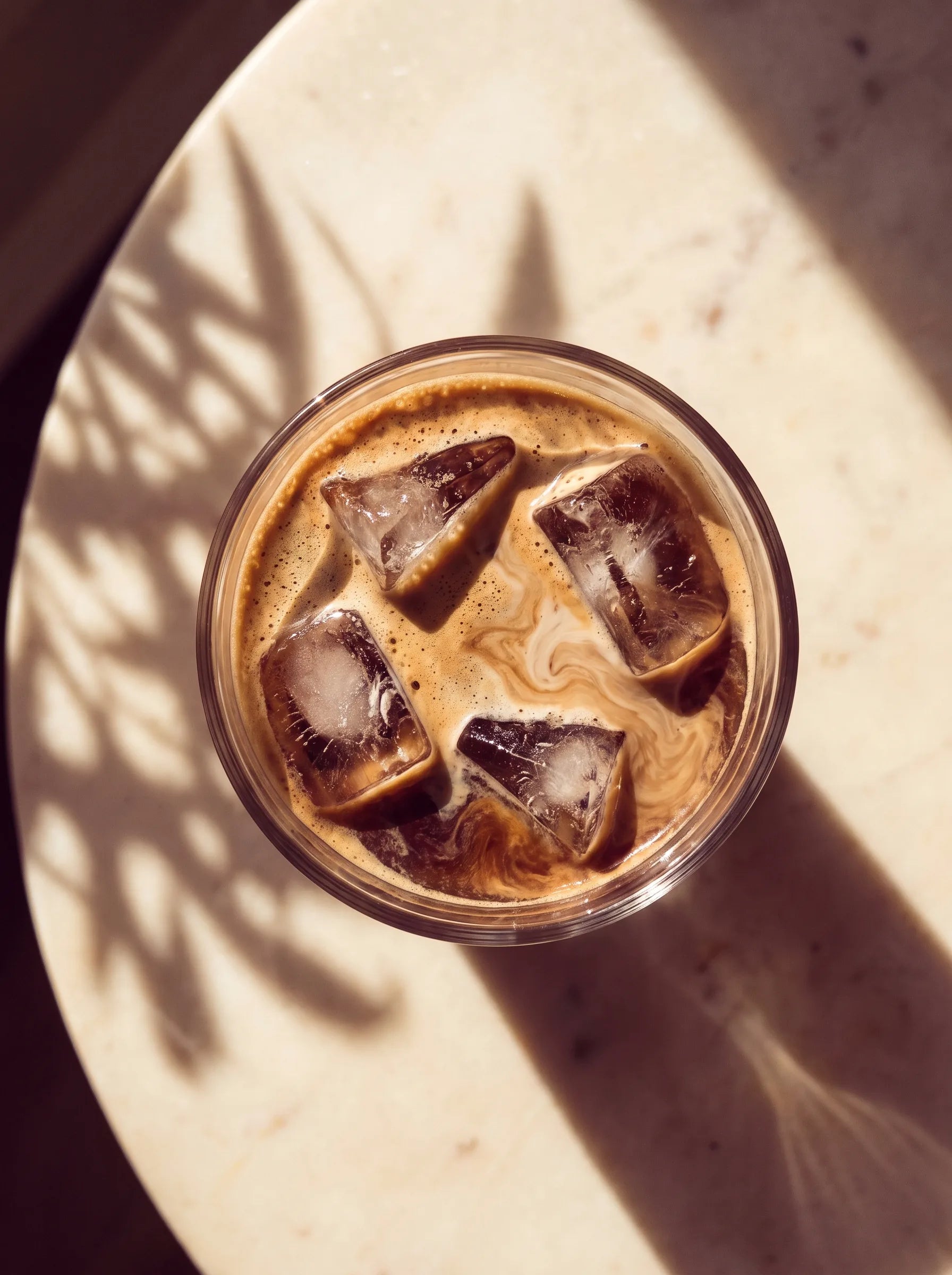 A top-down view of an iced coffee with cream swirling through the ice, on a sunlit stone surface.